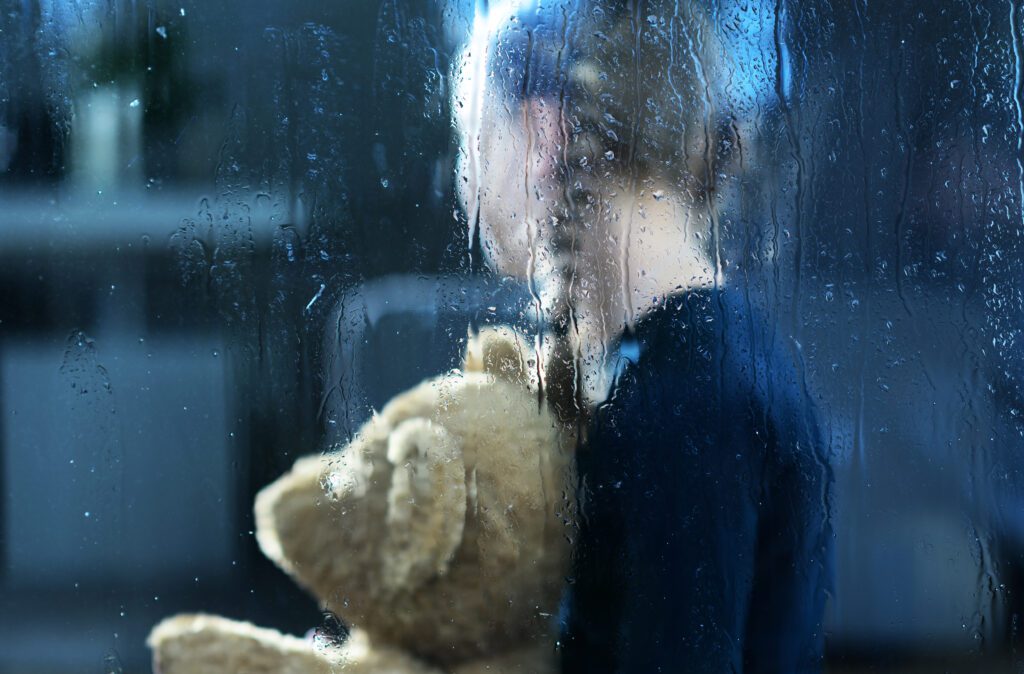 Kind hinter einer von Regentropfen bedeckten Scheibe mit Teddy im Arm, Rückansicht. Die Kehrseite der Schuldgefühle bei Ersatzkindern ist, dass sie selbst zu Tätern werden können, wenn sie keine andere Beziehungsform kennengelernt haben, als eine, die durch Schuld belastet ist.